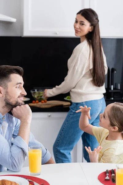 Gesto del niño mientras habla con papá sonriente y madre borrosa preparando el desayuno en la cocina - foto de stock