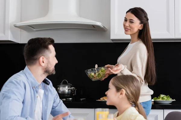 Cheerful woman holding bowl of fresh vegetable salad near blurred husband and daughter — Stock Photo