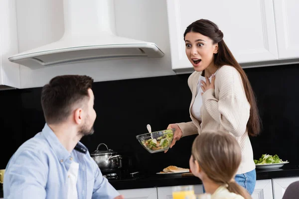 Donna stupita toccando petto mentre tenendo insalata di verdure vicino famiglia offuscata — Foto stock