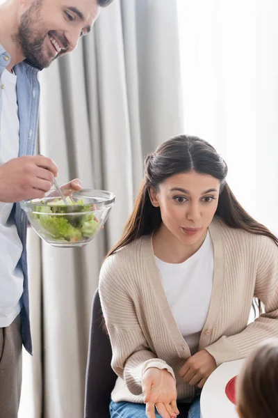 Mutter spricht mit verschwommenem Kind nahe glücklichem Ehemann mit Schüssel mit frischem Gemüsesalat — Stockfoto