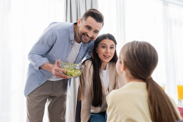 Visão traseira da criança borrada perto de mãe feliz e pai com tigela de salada de legumes — Fotografia de Stock