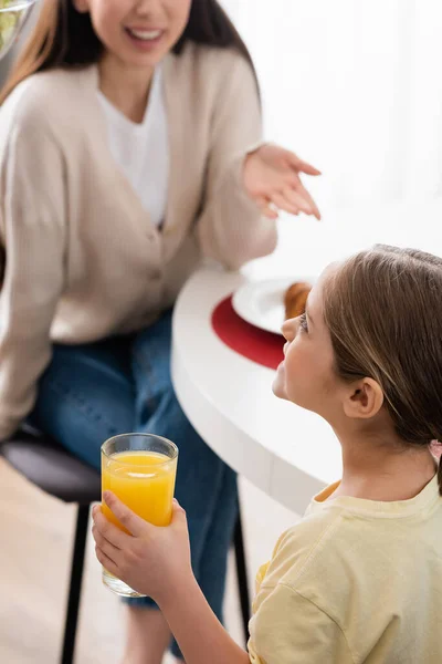 Joyeuse fille tenant verre de jus d'orange près de mère floue pointant avec la main — Photo de stock