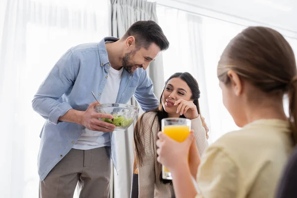 Homme heureux embrassant femme tout en tenant bol avec salade près de fille floue avec du jus d'orange — Photo de stock