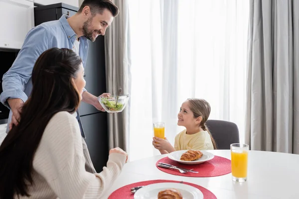 Homem alegre segurando tigela de salada de legumes perto de esposa e filha com copo de suco de laranja — Fotografia de Stock