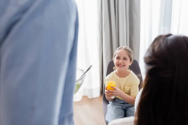 Alegre chica sosteniendo vaso de jugo de naranja cerca borrosa padres - foto de stock