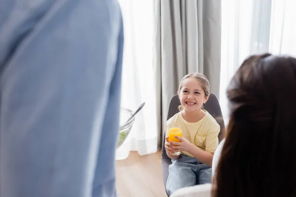 Menina alegre segurando vidro de suco de laranja perto de pais desfocados — Fotografia de Stock