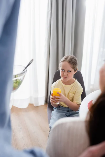 Displeased girl grimacing while holding orange juice near blurred father with vegetable salad — Stock Photo