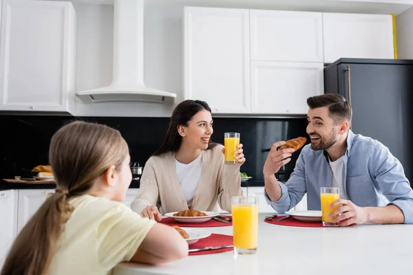 Homem alegre comendo croissant perto de esposa e filha em primeiro plano borrado — Fotografia de Stock