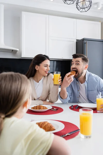 Веселый мужчина кусает круассан возле жены и размытая дочь во время завтрака — стоковое фото