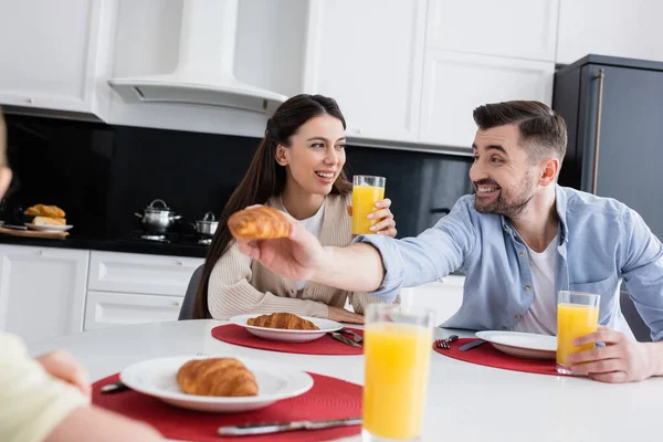 Веселый мужчина дает круассан размытой дочери рядом с улыбающейся женой во время завтрака — стоковое фото