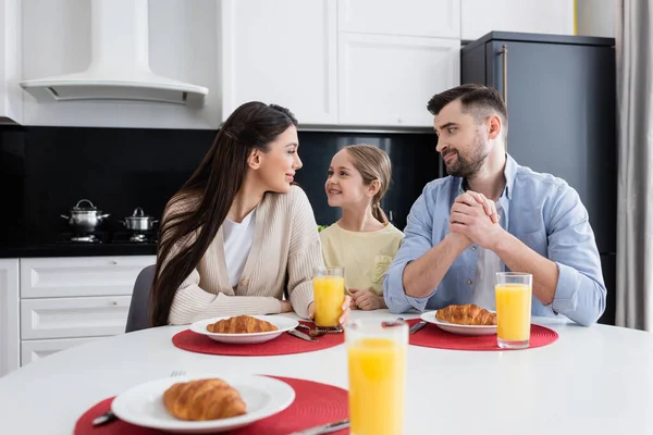 Familia feliz mirándose cerca de croissants y zumo de naranja en la cocina - foto de stock