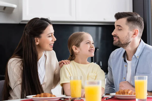 Ragazza gioiosa sorridente vicino a genitori felici, croissant e succo d'arancia in primo piano sfocato — Foto stock