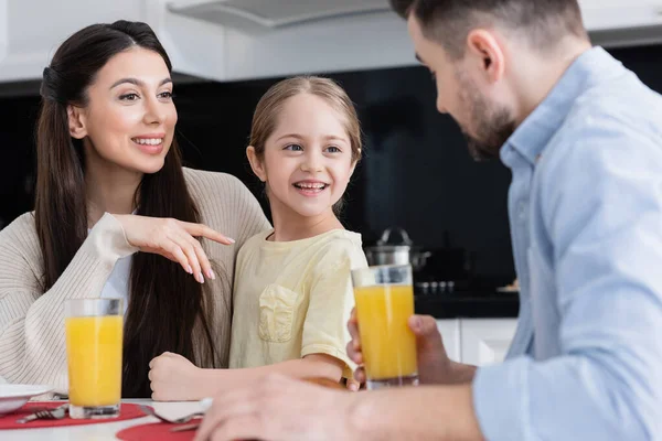 Glückliche Frau zeigt mit einem Glas Orangensaft auf fröhliche Tochter neben verschwommenem Ehemann — Stockfoto