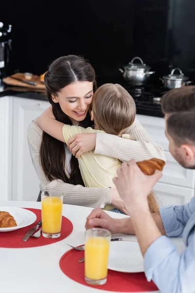 Fröhliche Frau umarmt Tochter in Küche neben verschwommenem Ehemann mit Croissant — Stockfoto