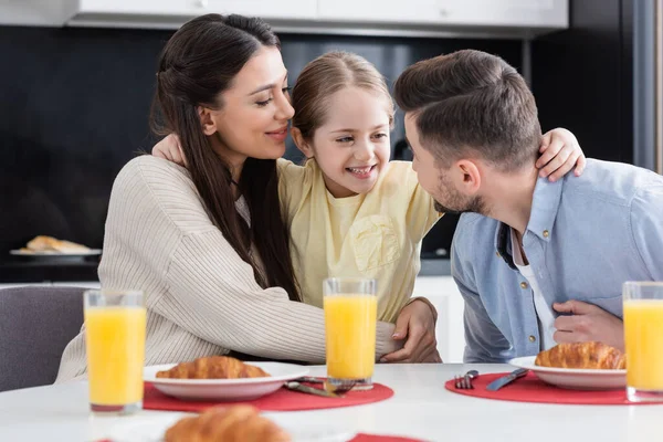 Menina feliz abraçando pais perto de croissants e suco de laranja na cozinha — Fotografia de Stock