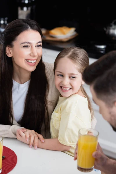 Alegre madre e hija tomados de la mano cerca borrosa papá celebración de jugo de naranja - foto de stock