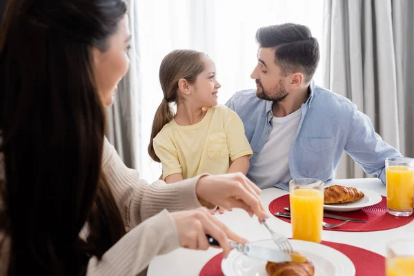 Mujer borrosa cortando croissant cerca feliz hija y marido mirándose durante el desayuno - foto de stock