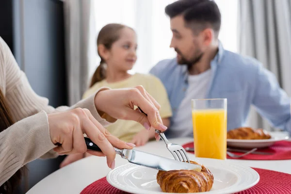 Frau schneidet leckeres Croissant in der Nähe von Mann und Tochter auf verschwommenem Hintergrund — Stockfoto