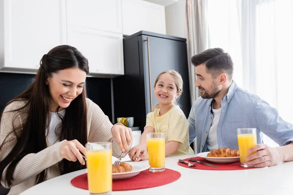 Cheerful woman cutting croissant during breakfast with husband and daughter — Stock Photo