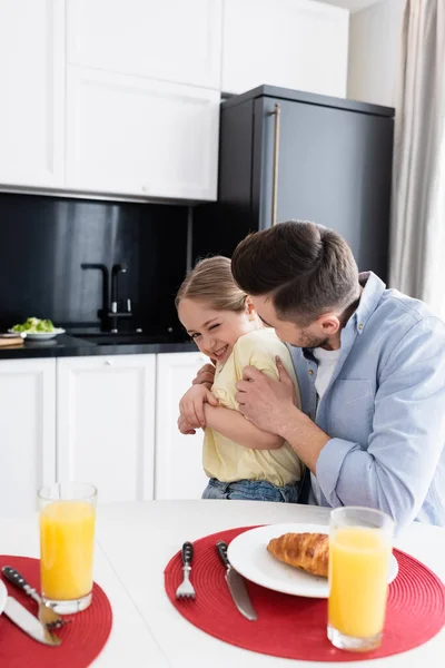 Padre abrazando alegre hija cerca de jugo de naranja y croissant en cocina - foto de stock