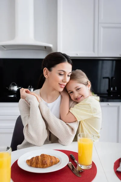 Улыбающаяся девушка обнимает счастливую мать возле круассана и апельсинового сока на кухне — стоковое фото