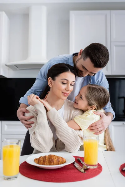 Счастливый мужчина обнимает жену и дочь во время завтрака на кухне — стоковое фото