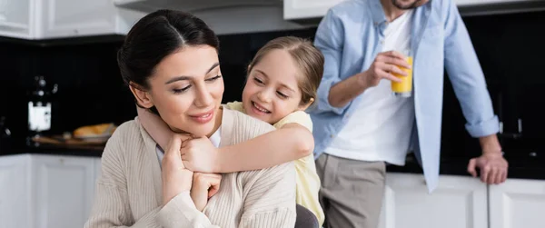 Sonriente chica abrazando feliz mamá cerca padre celebración de jugo de naranja sobre fondo borroso, bandera - foto de stock