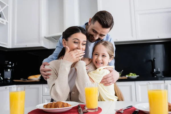 Mulher rindo cobrindo boca com a mão perto da filha e do marido durante o café da manhã — Fotografia de Stock