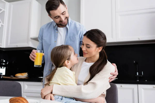 Mulher feliz abraçando filha perto do marido segurando suco de laranja na cozinha — Fotografia de Stock