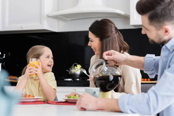 Размытый мужчина наливает кофе рядом с улыбающейся дочерью и женой разговаривая во время завтрака — стоковое фото