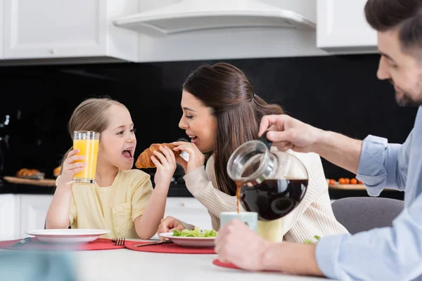 Verschwommener Mann schenkt Kaffee in der Nähe fröhlicher Frau ein, die Tochter mit Croissant füttert — Stockfoto