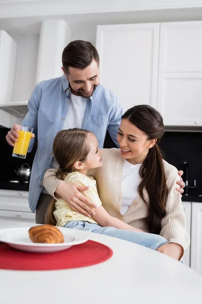 Donna gioiosa abbracciando figlia vicino al marito con succo d'arancia in cucina — Foto stock