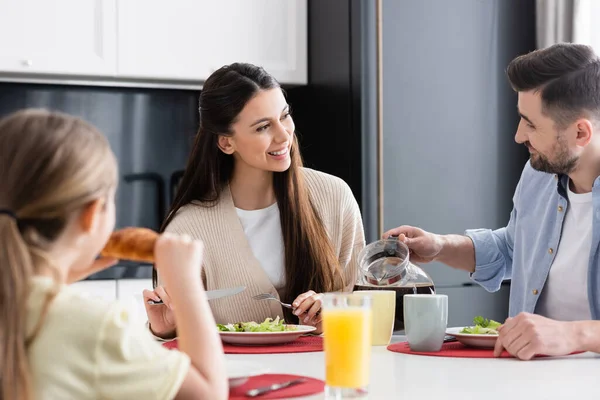 Uomo versando caffè vicino moglie gioiosa e figlia offuscata mangiare croissant per la prima colazione — Foto stock