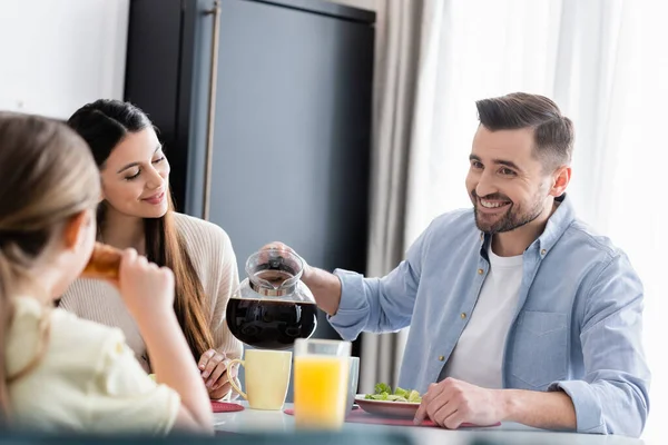 Sorrindo homem derramando café perto da esposa e filha borrada durante o café da manhã — Fotografia de Stock