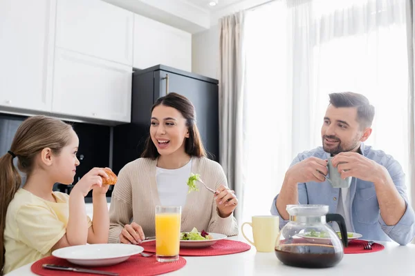 Mulher alegre comer salada de legumes para o café da manhã perto da esposa e do marido — Fotografia de Stock