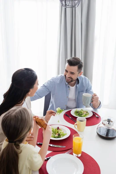 Feliz hombre sonriendo cerca de esposa e hija durante el desayuno - foto de stock