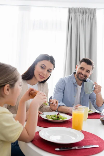 Ragazza mangiare croissant vicino genitori felici durante la colazione — Foto stock