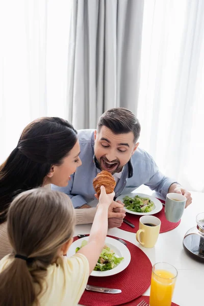 Mädchen füttert Vater beim Frühstück mit Croissant neben glücklicher Mutter — Stockfoto