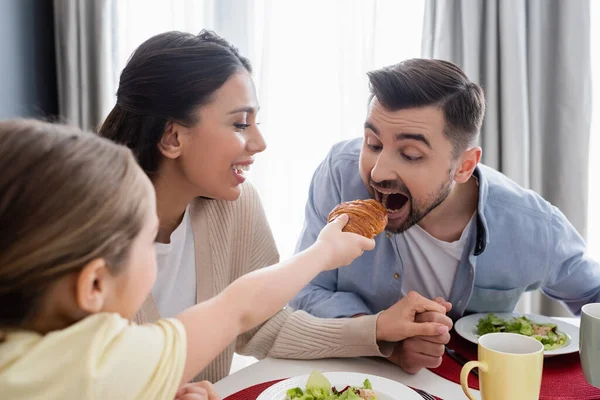 Blurred child feeding dad with croissant near cheerful mother during breakfast — Stock Photo