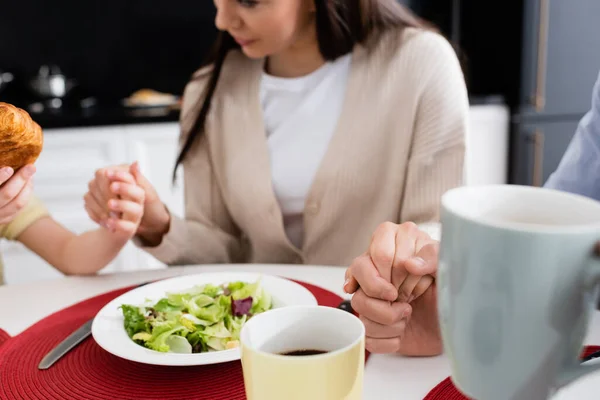 Cropped view of blurred woman holding hands of husband and daughter while praying before breakfast — Stock Photo