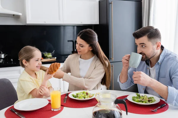 Allegra ragazza tenendo croissant vicino ai genitori e insalata di verdure durante la colazione — Foto stock