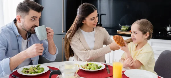 Frau flechtet Haare ihrer Tochter in der Nähe ihres Mannes, der während des Frühstücks Kaffee trinkt, Banner — Stockfoto