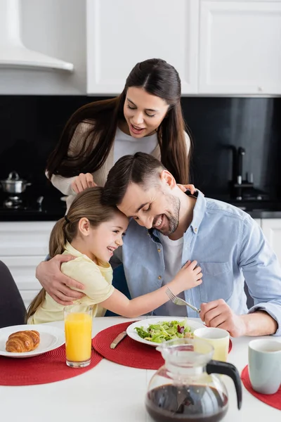 Улыбающаяся женщина, стоящая рядом с счастливыми мужем и дочерью, обнимающимися во время завтрака — стоковое фото