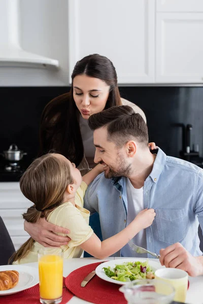 Woman pouting lips near daughter and father embracing during breakfast — Stock Photo