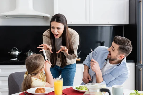Frau hat Spaß, während sie Tochter während des Frühstücks in der Nähe aufgeregter Ehemänner erschreckt — Stockfoto