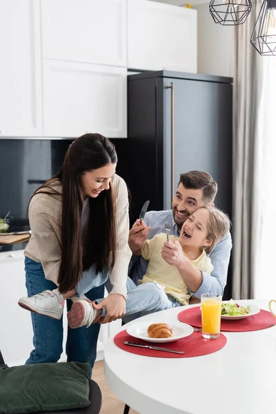 Aufgeregtes Mädchen hält Gabel und Messer in der Hand und amüsiert sich beim Frühstück mit den Eltern — Stockfoto