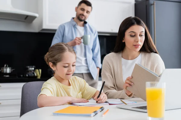 Woman reading book while helping daughter doing homework near blurred husband drinking coffee — Stock Photo