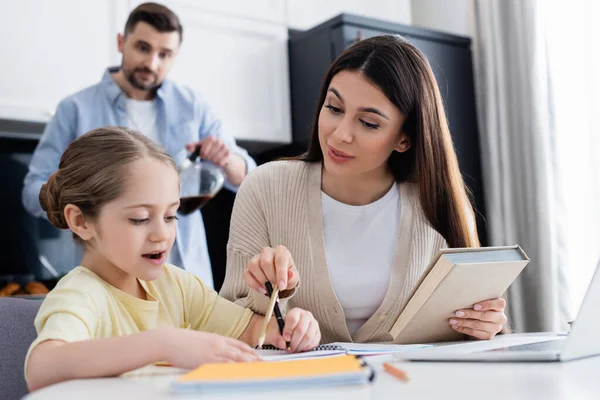 Femme pointant au crayon tout en aidant sa fille à faire ses devoirs près d'un mari flou — Stock Photo