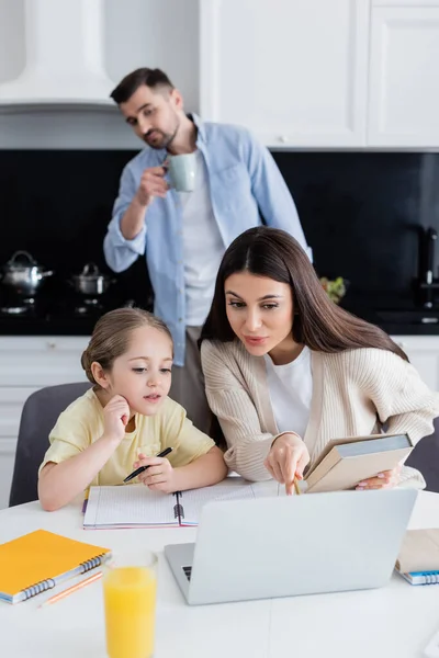 Femme pointant avec crayon à l'ordinateur portable tout en aidant fille dans les devoirs près de mari flou — Photo de stock