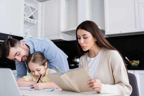 Mulher lendo livro perto da filha escrevendo no caderno e sorrindo marido — Fotografia de Stock